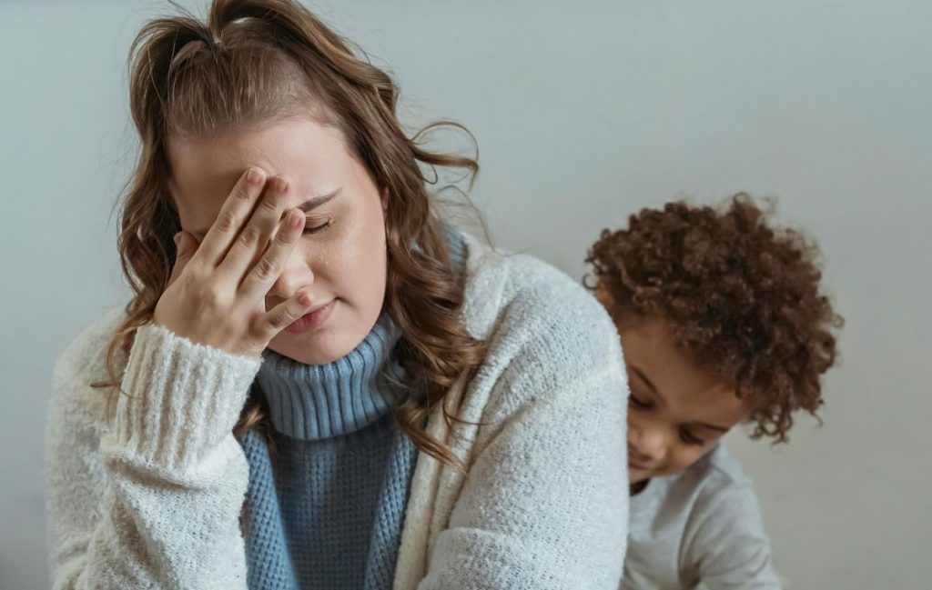 Exhausted mother sitting on a couch with her young son, resting her head on her hand while the child leans on her, conveying parental fatigue and emotional overwhelm.